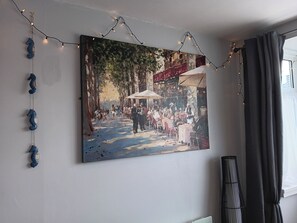 Una pintura de una escena callejera con gente y edificios, colgada en una pared con luces colgantes.