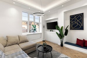 A modern living room with a sectional sofa, a round coffee table, and a large window with a view of buildings. - Apartment - Sleeps 8 - Golders Green 10 mins (London)