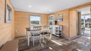 Un intérieur moderne avec des murs en bois, un coin repas avec des chaises blanches, une cuisine avec des meubles blancs et une grande fenêtre offrant une vue dégagée.