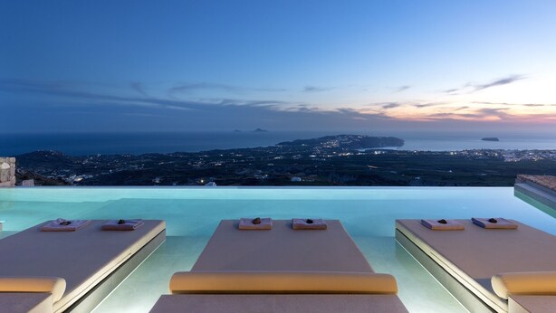 Una piscina infinita con sillas de descanso y vistas a un paisaje costero al atardecer.