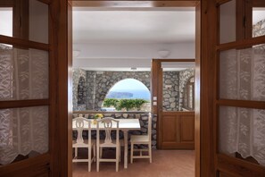 A view through a wooden-framed doorway into a room with a stone wall, a white table, and chairs. - One bdr Villa-Sleeps 4-Hot Tub-Sea View-Parking (Pyrgos)