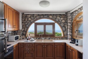 A kitchen with wooden cabinets, a stone wall, a built-in oven, a microwave, and a large window with a view of the ocean. - One bdr Villa-Sleeps 4-Hot Tub-Sea View-Parking (Pyrgos)