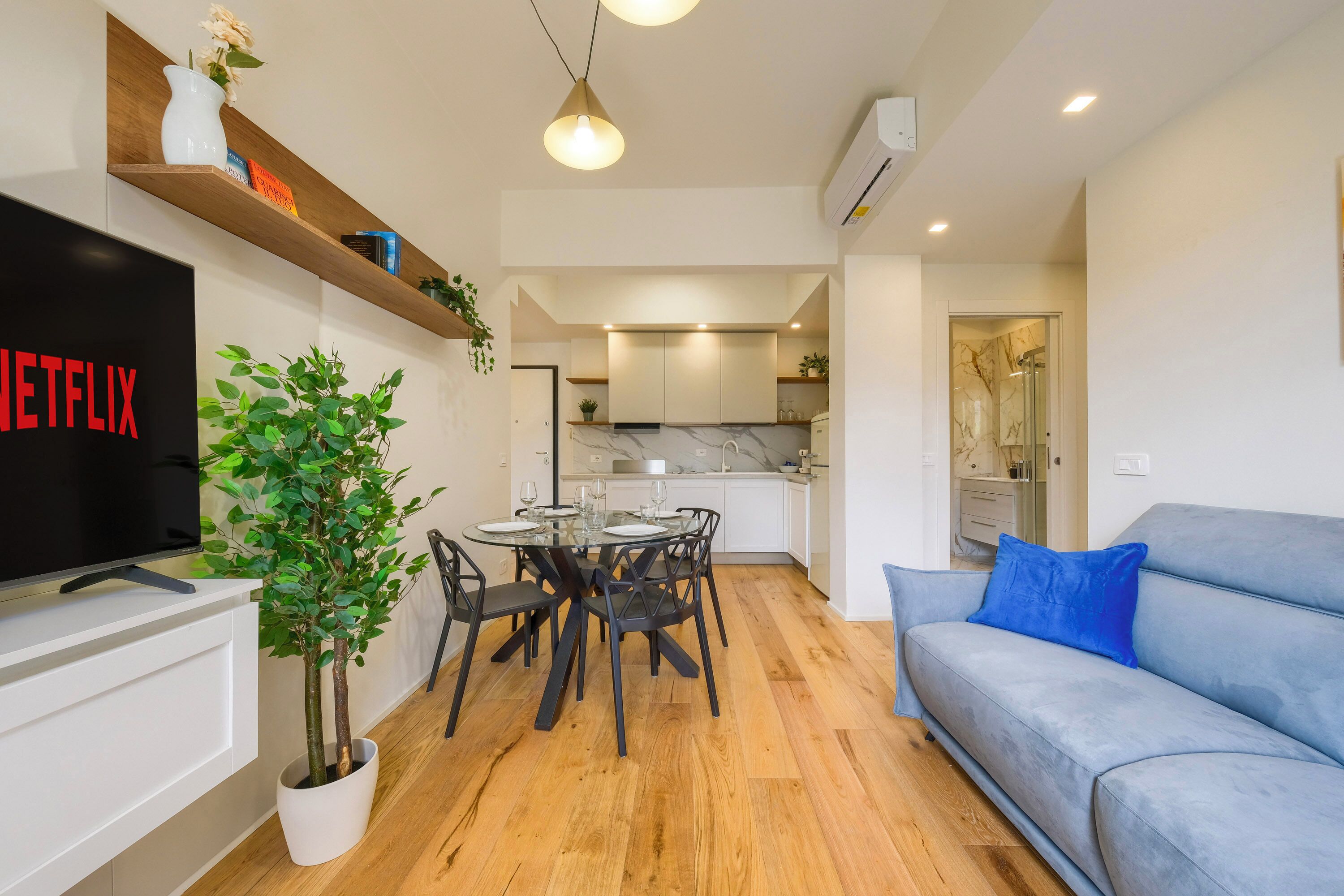 A modern living room with a flat-screen TV, a dining table with chairs, a blue sofa, and a potted plant.