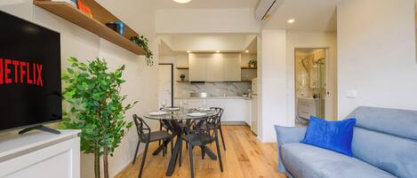 A modern living room with a flat-screen TV, a dining table with chairs, a blue sofa, and a potted plant.