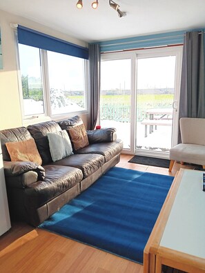 Ein Wohnzimmer mit braunem Ledersofa, einem blauen Teppich, einem Holztisch und Blick auf einen Garten durch eine Schiebetür.