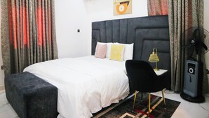 Une chambre bien rangée, avec un lit, un bureau, une chaise, un climatiseur, un ventilateur et une œuvre d’art accrochée au mur.