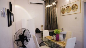 Un espace repas moderne avec une table en verre, des chaises blanches, un ventilateur noir, une plante en pot et un climatiseur fixé au mur.