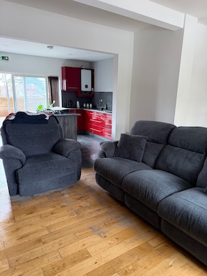 A modern living room with a grey sofa and armchair, wooden flooring, and a red kitchen in the background. - Charming Birmingham 3 Bedroom Apartment (Birmingham)