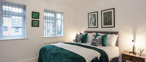 A bedroom with a bed, two windows, a radiator, and wall-mounted artwork.