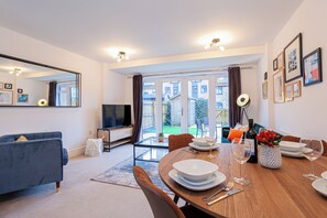 A modern living room with a sofa, a TV, a dining table set for two, and a view of the outside.