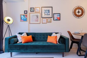 Un canapé de couleur bleu-vert avec des coussins orange, un lampadaire et un mur orné de tableaux encadrés.