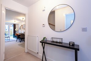 A hallway with a round mirror, a radiator, a console table with a plant and candles, and a dining area visible through an open door.