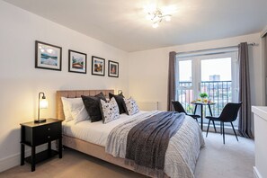 A bedroom with a bed, bedside table, lamp, framed pictures, and a balcony with a table and chairs.