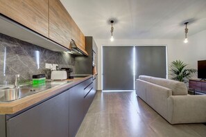 A modern kitchen with a marble backsplash, wooden cabinets, and a built-in oven. There is a dining area with a sofa and a TV. - Kuya Penthouse Suite / Heated Pool / City View (Swieqi)