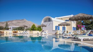 Una piscina azul cristalina con sillas de descanso y sombrillas, rodeada de edificios blancos con detalles azules y una montaña al fondo.