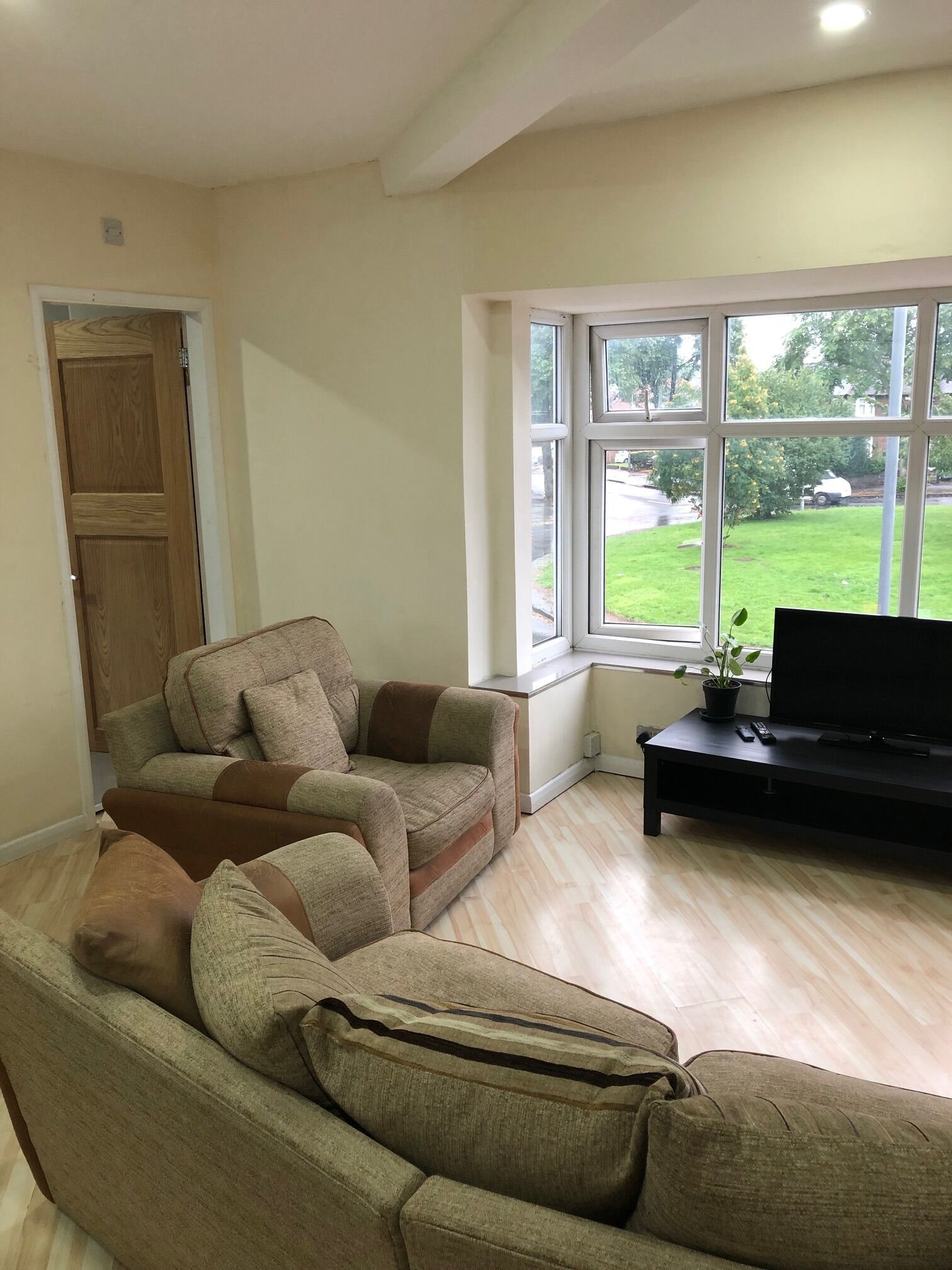 Una sala de estar con un sofá color beige, un mueble de televisión negro y una ventana con vista a un césped verde y árboles.