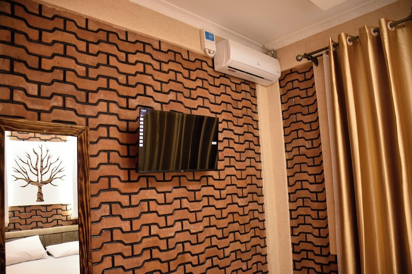 A room with a brick-patterned wall, a mounted air conditioner, a bed with white bedding, and a window with a tree decoration.
