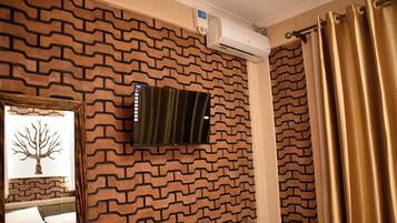 A room with a brick-patterned wall, a mounted air conditioner, a bed with white bedding, and a window with a tree decoration.