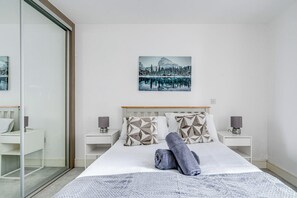A modern hotel room with a large bed, two bedside tables, a glass partition, and a painting of a snowy landscape. - Battersea 2-Bed Apt, Sleeps 4 with WiFi (London)