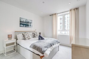 A bedroom with a bed, a nightstand, a lamp, a painting, and a window with curtains. - Battersea 2-Bed Apt, Sleeps 4 with WiFi (London)