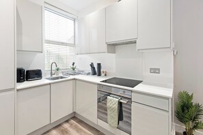 A modern kitchen with white cabinetry, a black induction cooktop, a stainless steel sink, and a window with blinds. - Apartment - Sleeps 11 - Hampstead HighStreet 5mins (London)