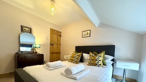 A neatly made bed with white linens and rolled towels, a wooden nightstand with a lamp, a bedside table with a lamp, and a framed picture on the wall. - Ulverston Cozy Cottage Retreat (Ulverston)