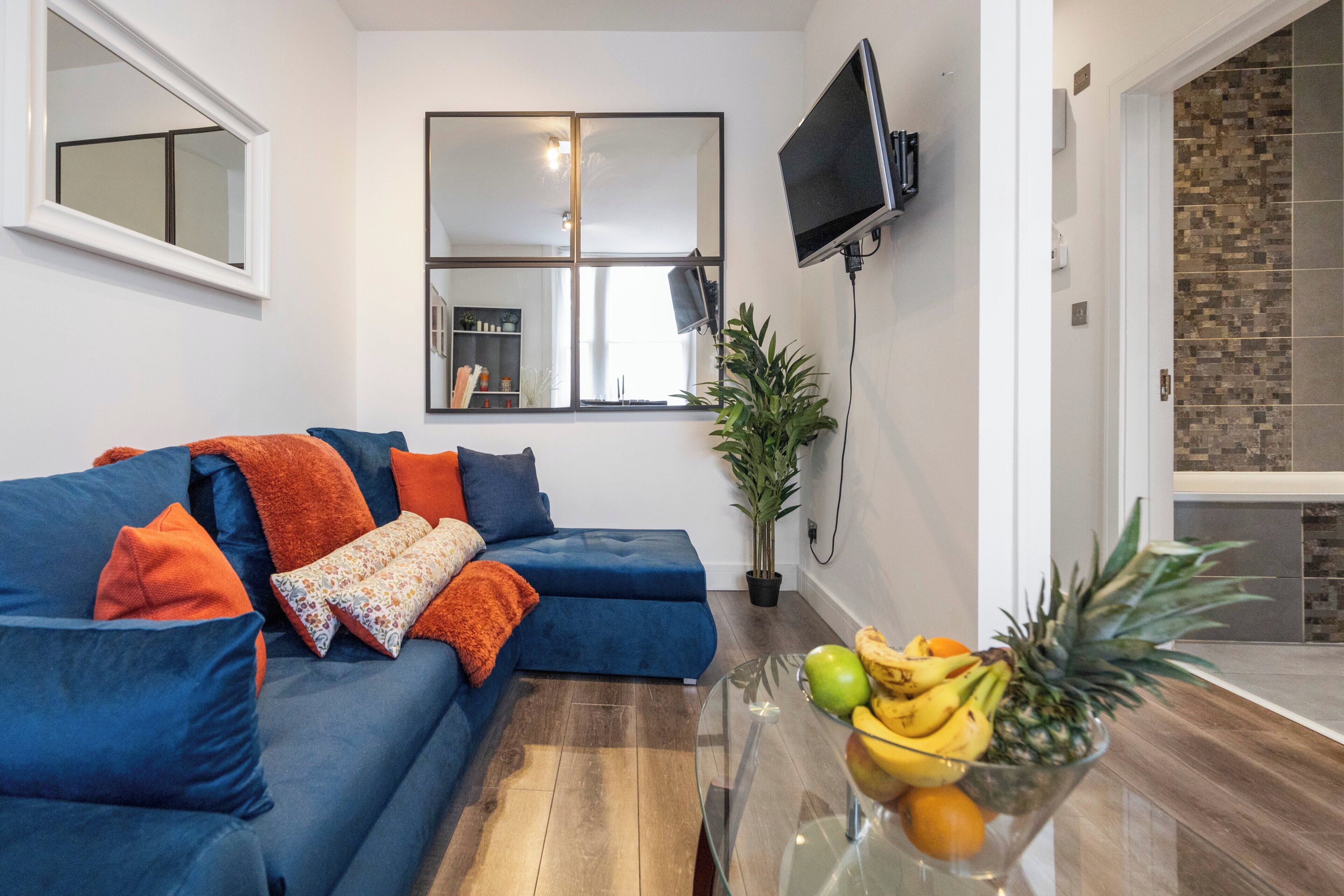 A living room with a blue sofa, a glass coffee table with fruit, a TV mounted on the wall, and a potted plant.