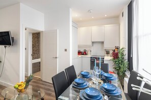 A modern dining area with a glass table set for four, a kitchen in the background, and a TV mounted on the wall. - Charming London Apartment in Central London (London)