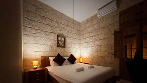 Ett sovrum med en vägg som har en stenliknande struktur, en säng med kuddar, ett sängbord med en lampa och en dörr av trä.