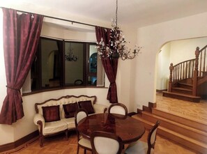 A dining area with a round wooden table and chairs, a sofa with cushions, a chandelier, and a staircase leading to an upper floor.