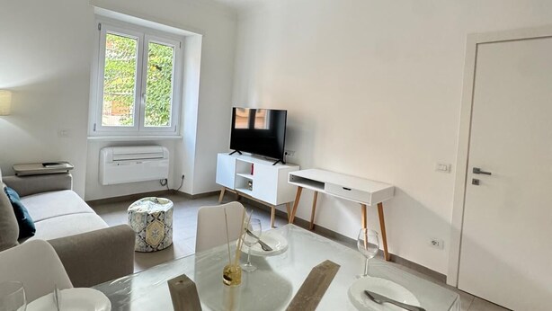 A modern living room with a glass dining table, white sofas, a flat-screen TV, and a large window with a view of greenery. - King of Rome Metro Hub - Rome Collection Metro A (RE DI ROMA) 15 dalColosseo (Roma)