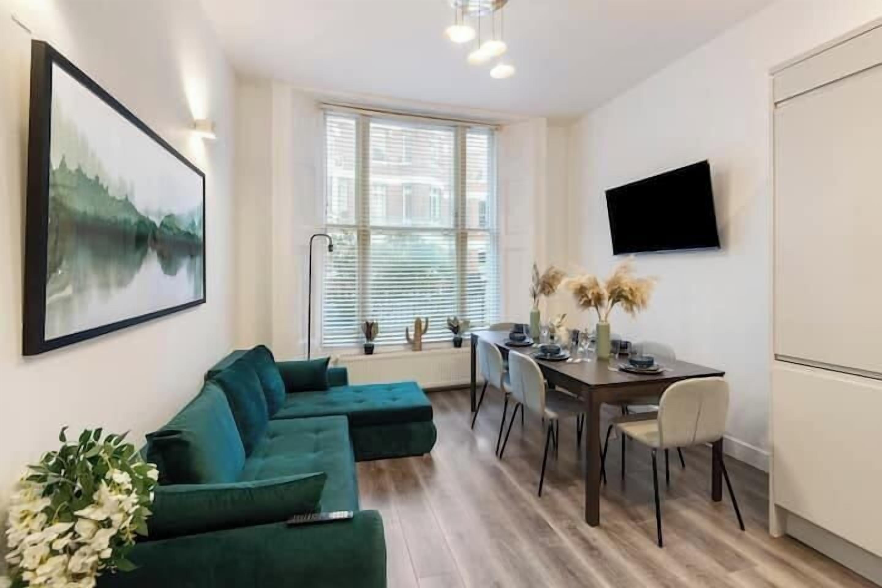 Een moderne woonkamer met een hoekbank, een eettafel met stoelen, een flatscreen televisie aan de muur en een raam met zonwering.