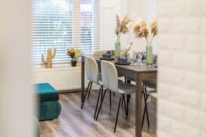 A dining area with a table set for four, chairs, a window with blinds, and a radiator. - Apartment - Sleeps 7 - Paddington Station 5 mins (London)