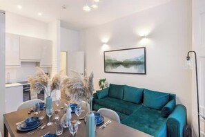 A modern living room with a teal sofa, a dining table set with blue plates and glasses, and a painting of a landscape on the wall. - Apartment - Sleeps 7 - Paddington Station 5 mins (London)