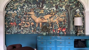 Una stanza con un murale raffigurante un cervo e vegetazione, un comò blu con cassetti e una lampada su un tavolino.