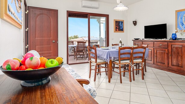 A dining area with a table, chairs, a bowl of fruit, a television, and a view through a glass door. - Holiday Home 'Trilo Stazzu' with Private Terrace, Private Garden and Air Conditioning (San Teodoro)