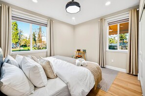 Ein modernes Hotelzimmer mit einem großen Bett, zwei Fenstern mit Vorhängen, einem kleinen Tisch mit einem Stuhl und Blick auf einen Park.