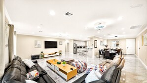 A spacious living room with a leather sofa, a wooden coffee table, and a TV. There is a kitchen in the background with white cabinets and a dining area with a table and chairs. - Spacious 9BR Home + Parking + Near DTLA #TravelSGV (Avocado Heights)