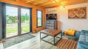 A cozy living room with a wooden ceiling, a blue sofa, a television, and a sliding glass door leading to a garden.