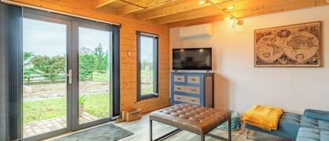A cozy living room with a wooden ceiling, a blue sofa, a television, and a sliding glass door leading to a garden.