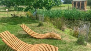 Two wooden loungers in a grassy area with a pond and a wooden structure in the background.