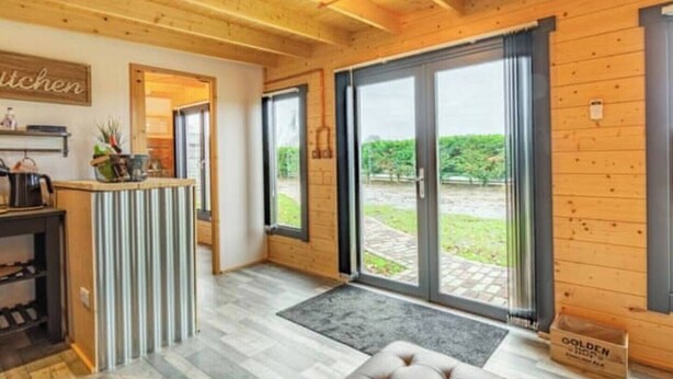 A wooden interior with a kitchen area, a seating area with a tufted ottoman, and a view of a garden through large sliding glass doors.