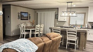 Una sala de estar moderna con un sofá seccional marrón, comedor con sillas blancas y una cocina con encimera de madera.