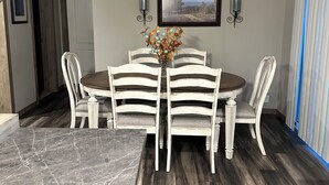 Un comedor con una mesa redonda y seis sillas, un jarrón con flores, dos apliques de pared y un cuadro en la pared.