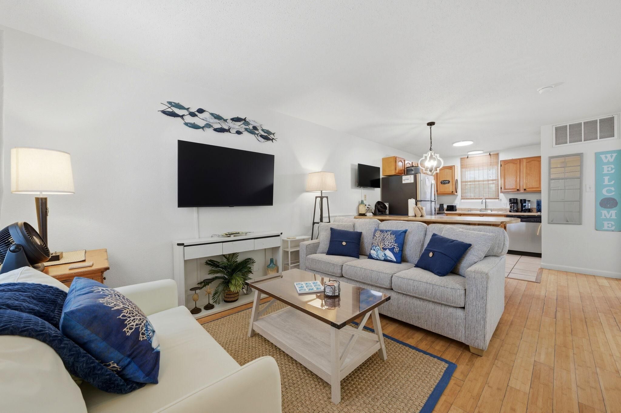Una sala de estar moderna con sofá, mesa de centro y televisor. Se aprecia una cocina al fondo con armarios de madera y una zona de comedor.