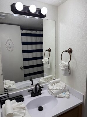 A bathroom with a white sink, black faucet, and a mirror with lights. There are towels on the counter and a striped shower curtain in the background. - Sunrise Village 118 (Gulf Shores)
