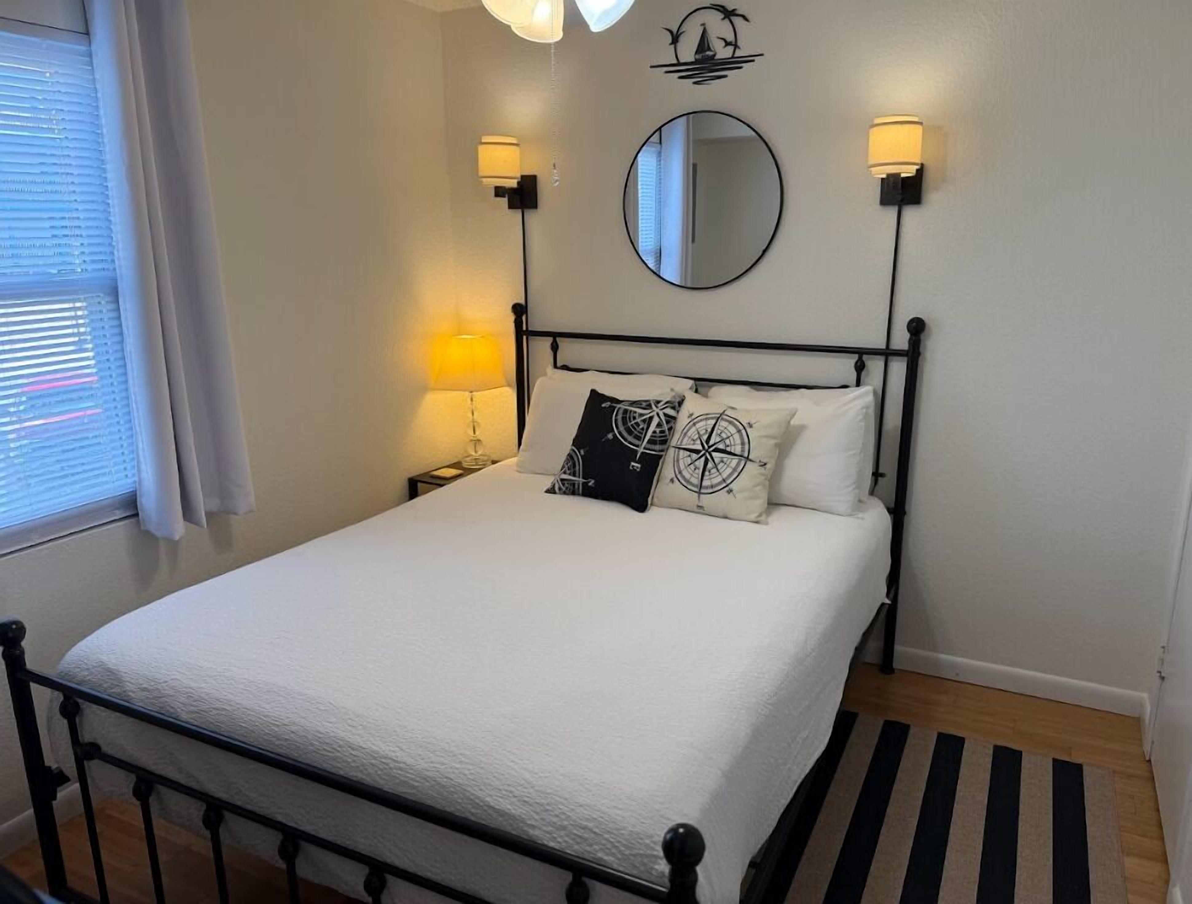 Una cama bien hecha con una colcha blanca y dos almohadas, una mesita de noche con una lámpara, un espejo redondo en la pared y una alfombra a rayas en el suelo.