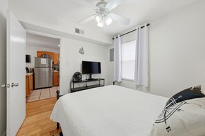 A bedroom with a bed, a ceiling fan, a TV on a stand, a window with curtains, and a kitchen in the background. - Sunrise Village 118 (Gulf Shores)