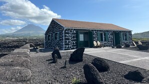 A stone building with a tiled roof, green shutters, and a paved entrance area with large rocks.