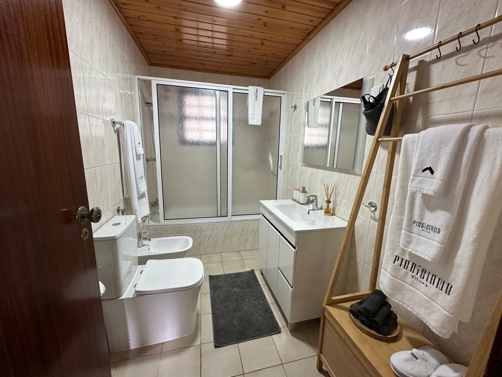 Un baño moderno con ducha, inodoro, lavabo y un soporte de madera con toallas y pantuflas.